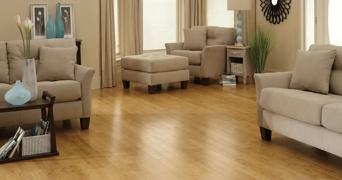 A living room with professionally installed bamboo flooring, beige sofas, a matching ottoman, a side table with a blue lamp, vases, framed pictures, and large windows with blinds and curtains in West Palm Beach, FL